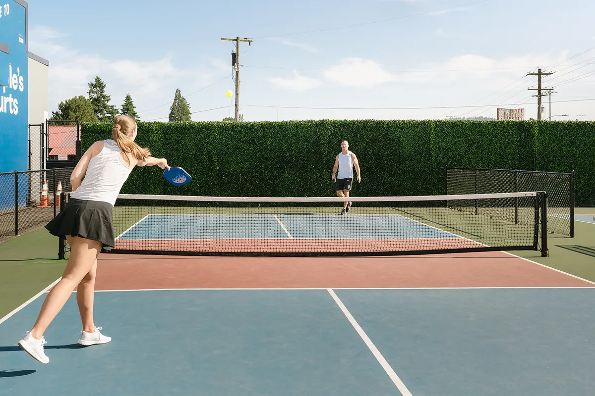 Two people play pickleball on an outdoor court. The woman in a black skirt hits a ball. Blue sky and hedge wall in the background.