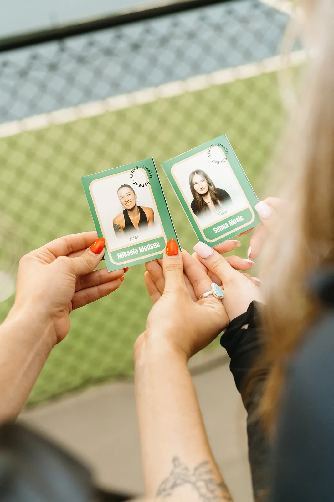 Two hands holding green cards with photos and names, Mikaela Bledsoe and Selma Music, against a blurred tennis court background.