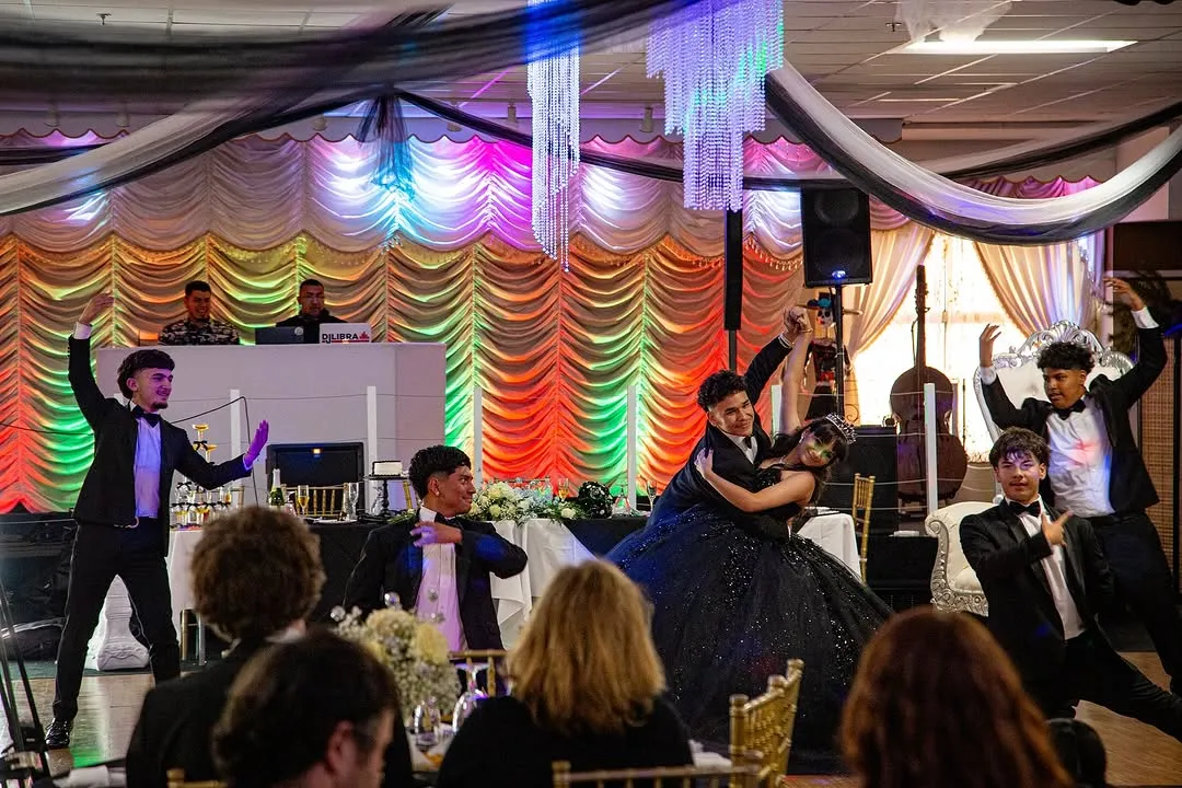Young men in tuxedos perform a dance around a couple in a ballroom, with colorful drapes and chandeliers. DJ booth and guests in background.