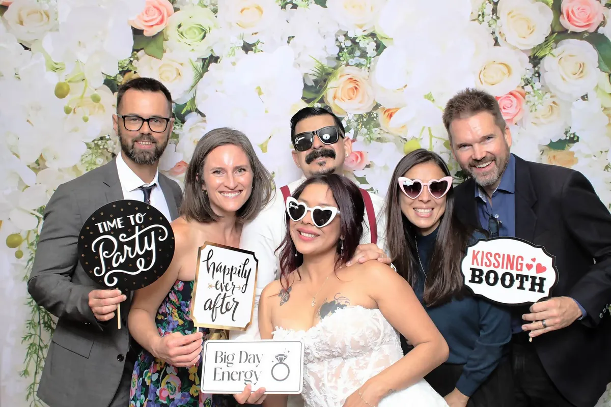 Group of people smiling, holding signs like "Time to Party" and "Kissing Booth." Background of white and pink roses. Celebratory mood.